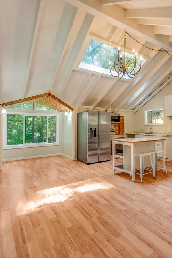A spacious, bright kitchen with light wood flooring, a large window showing green trees, a stainless steel refrigerator, a small kitchen island with two stools, and a chandelier hanging from the ceiling with skylight and sloped roof.