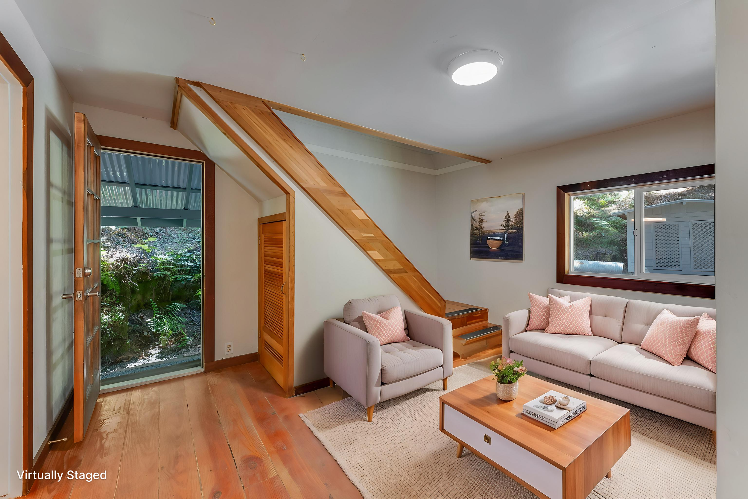 Living room with beige sofa and armchair, wooden coffee table with plants and books, window with view of trees outside, artwork on the wall, and an open door leading outside.