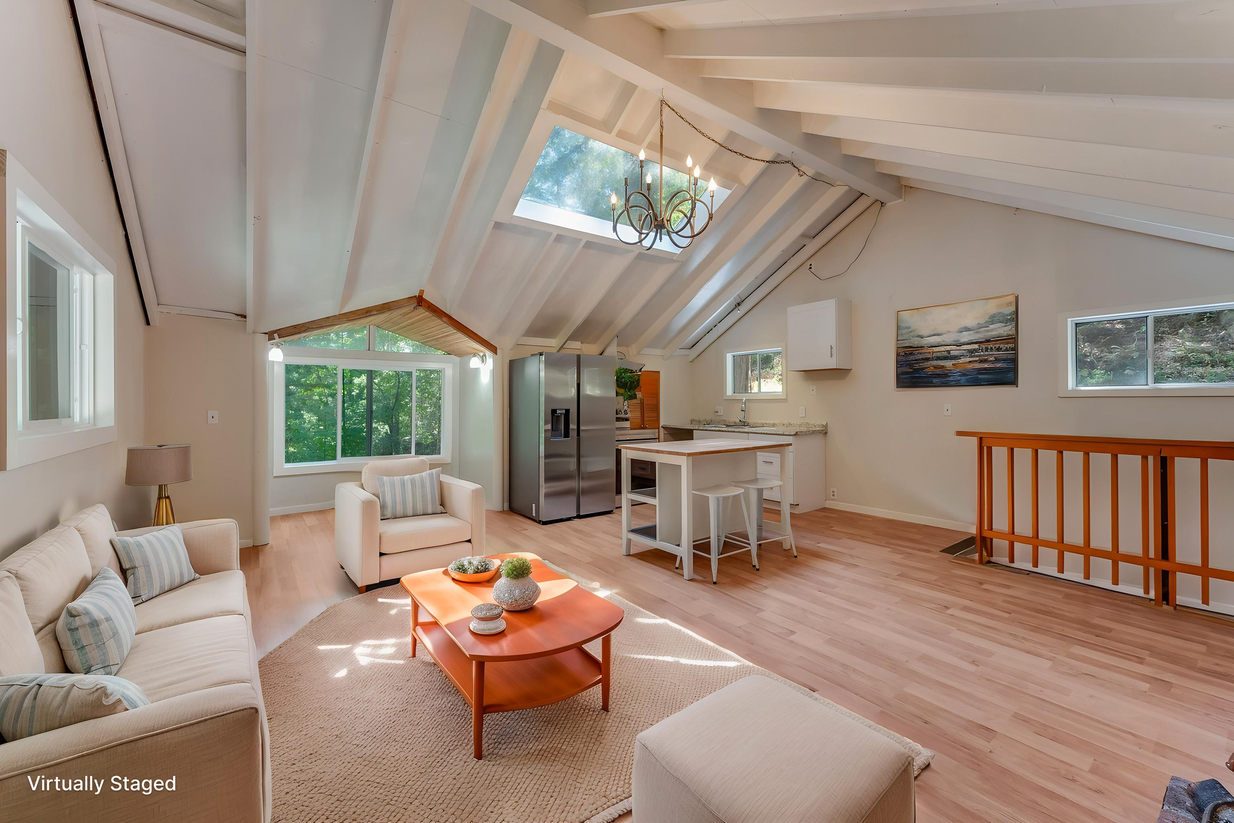 Living room with cream-colored sofa, armchair, orange coffee table, light wood flooring, large windows, skylight, chandelier, artwork on the wall, kitchen area with stainless steel refrigerator, white bar stools, and kitchen counter.