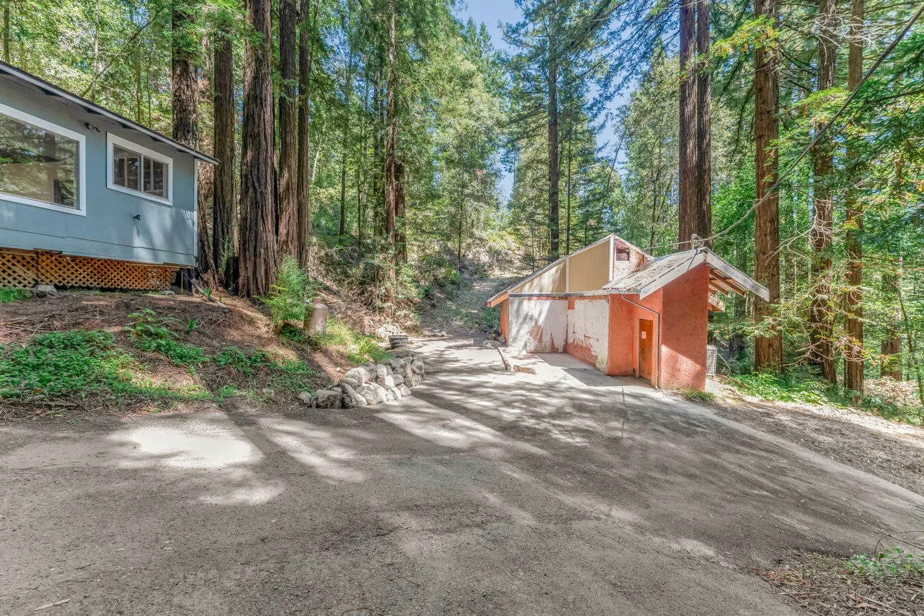 A dirt driveway in a wooded area with tall trees surrounds a partially constructed or renovated small building with orange and white walls and a sloped roof, and a second house with blue siding on the left.