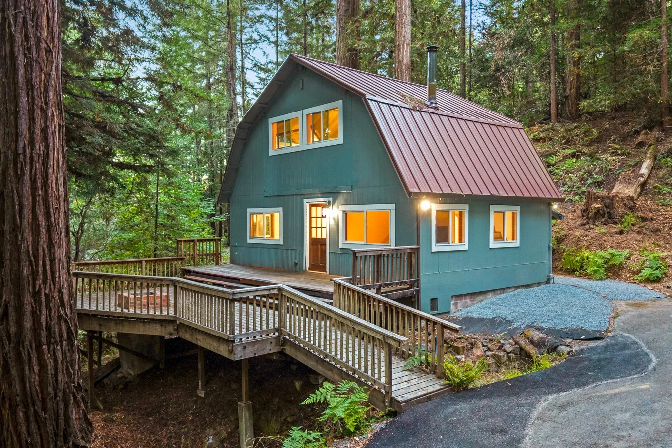 A blue two-story house with a red metal roof and white window frames, situated in a forested area. The house has a wooden deck and a ramp leading to the front door, with warm lights glowing from the windows.