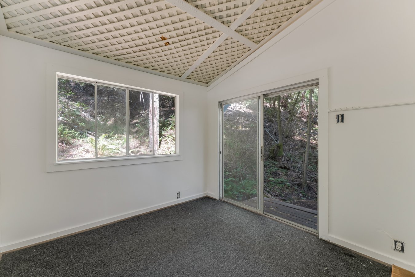 Empty room with white walls, a window, and sliding glass door leading outside to a wooded area.