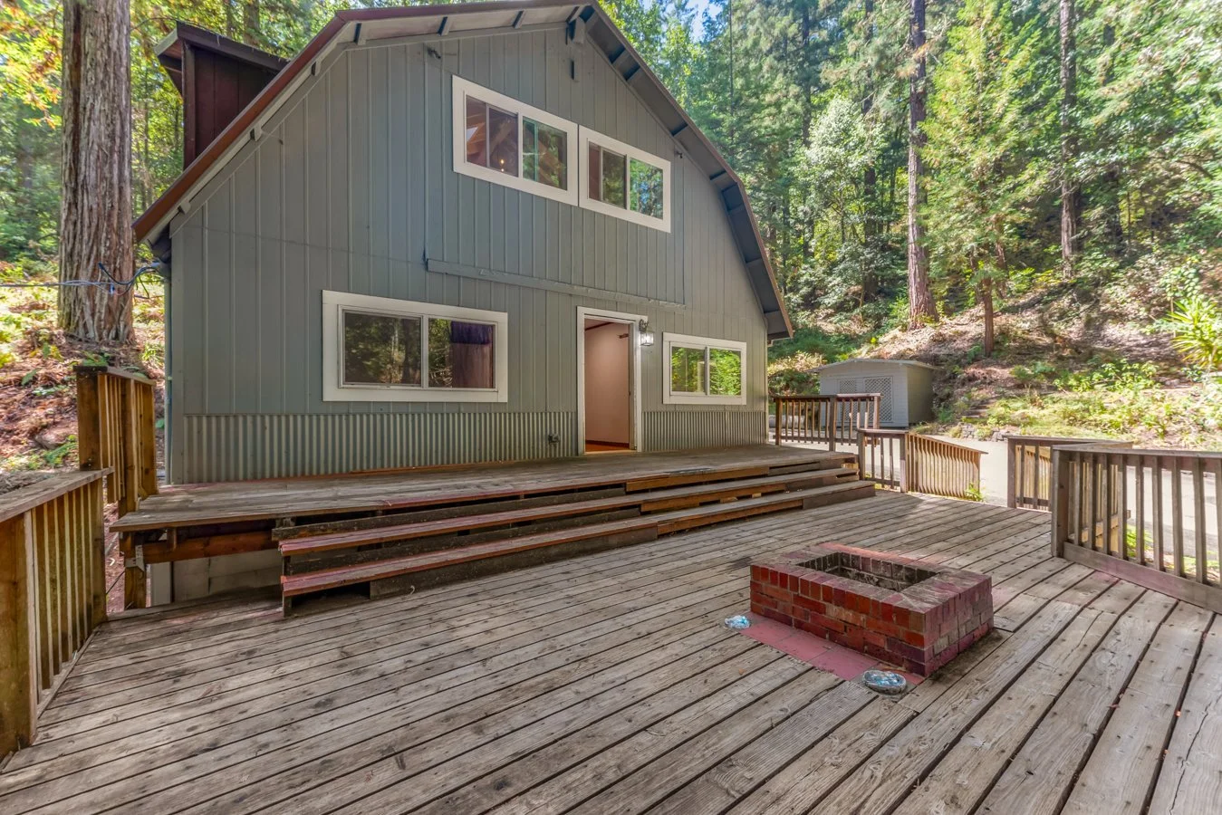 Backyard deck with stairs, a brick firepit, and a house with gray siding and multiple windows, surrounded by a forest.
