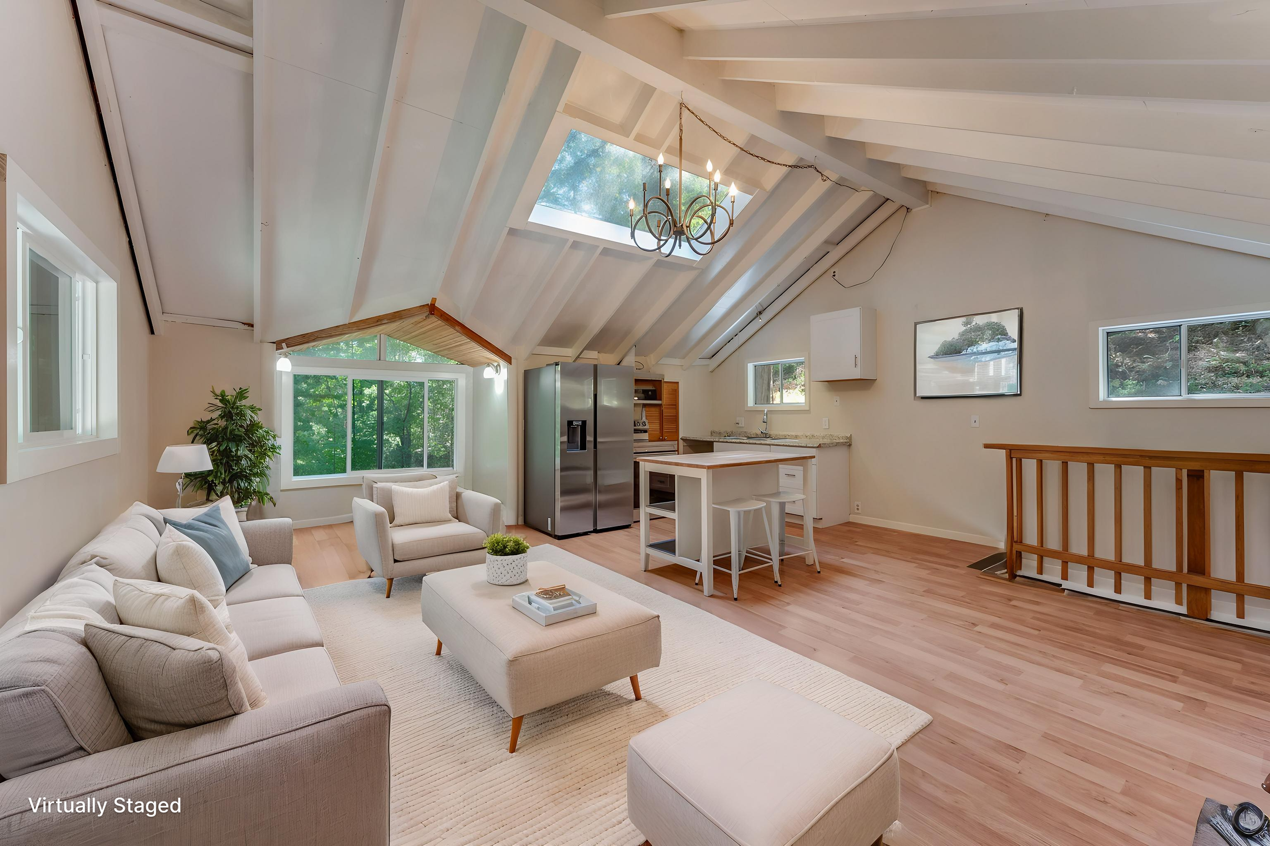 Living room with white sofa and chairs, a beige ottoman, large window with greenery outside, wood floor, ceiling with skylight, chandelier, kitchen area with stainless steel refrigerator and wooden cabinets, staircase with wooden railing, and a telev