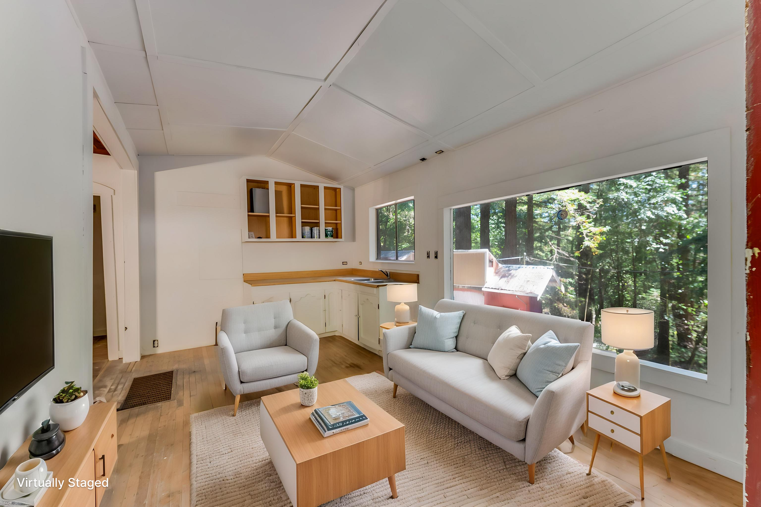 Living room with white sofa and armchairs, wooden coffee table with magazines, side table with lamp, large window showing trees outside, loft kitchen with open shelves, ceiling with angled panels, hardwood floor, and a red brick wall on the right.