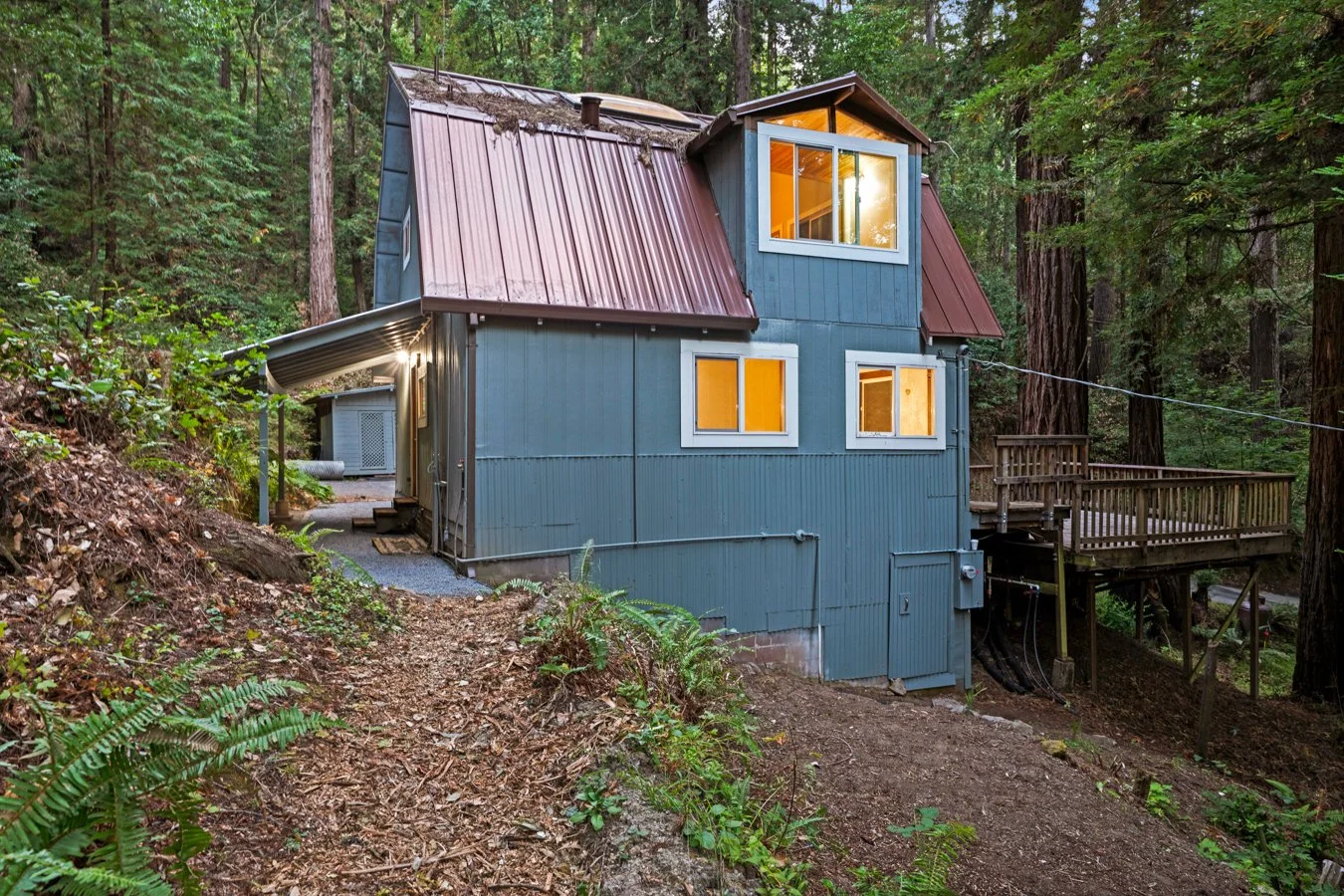 A two-story house with blue siding and a metal roof, nestled in a wooded area surrounded by tall trees, with warm lights glowing from the windows.