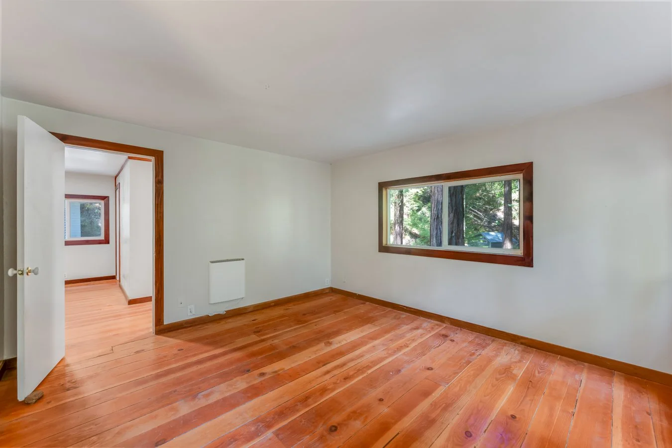 Empty room with wooden floors and a large window overlooking trees.