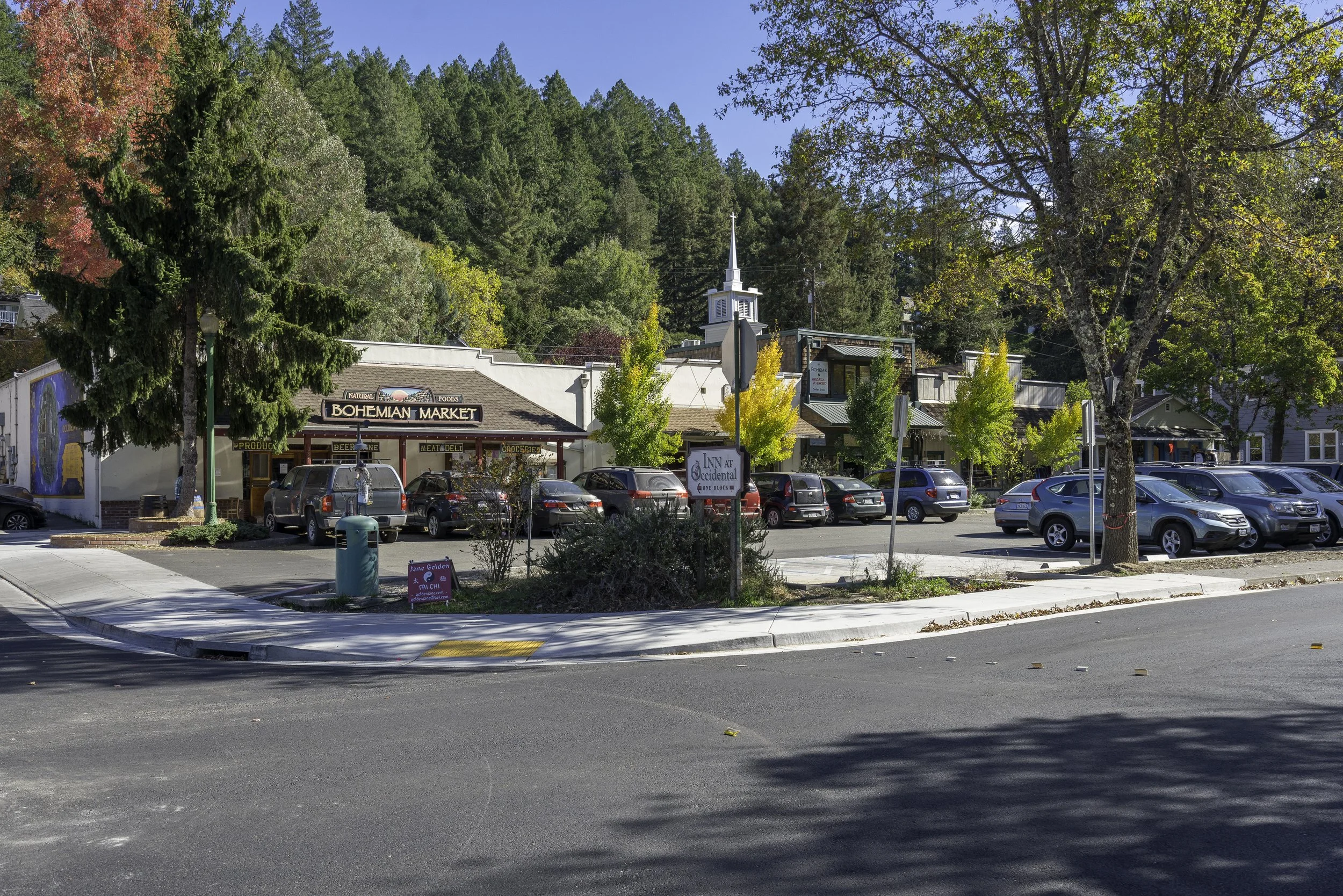 A small town street with a parking lot in the foreground, surrounded by trees with fall foliage. In the background are buildings including a market and other shops, with a hillside covered in dense trees behind them.