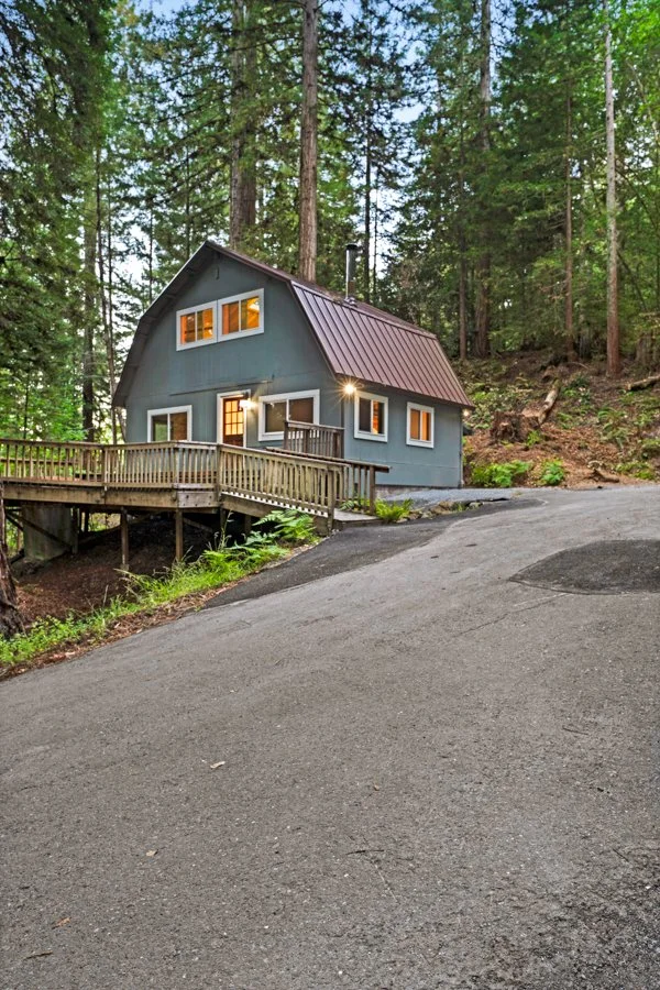 A gray house with a steep metal roof and multiple windows, surrounded by tall trees in a forested area. The house has a wooden deck with railing and is located at the end of a paved driveway.