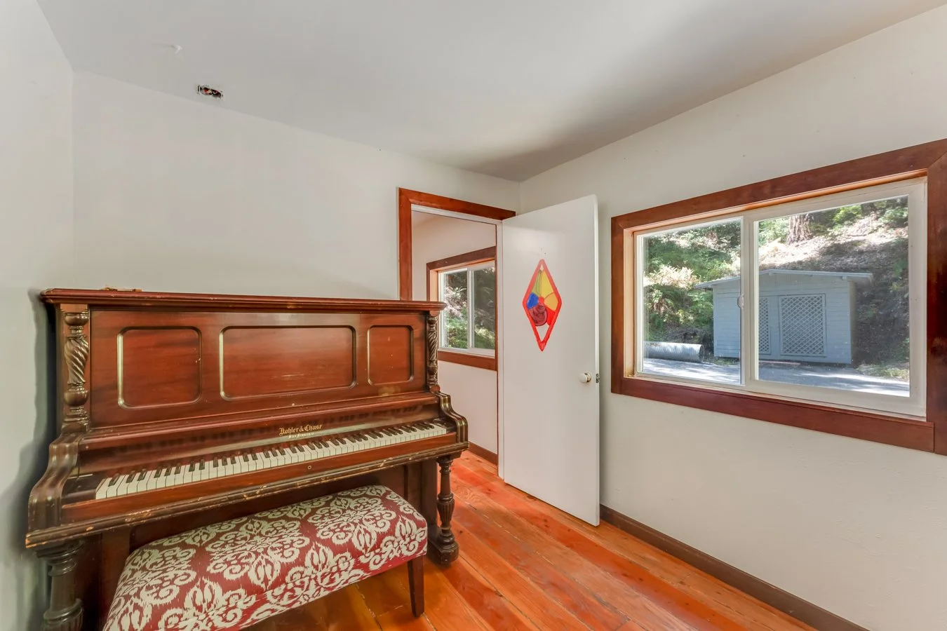 A room with a wooden upright piano and a cushioned bench, two windows with wooden frames, a white door with a colorful diamond-shaped decoration, and hardwood floors.