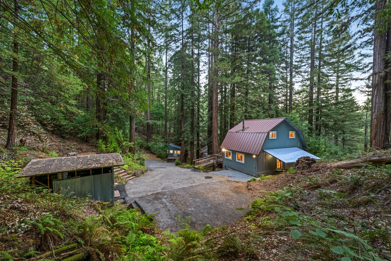 A cozy house in a dense forest with tall trees, showing a gravel driveway, a small covered shed, and warm lights glowing from the windows.