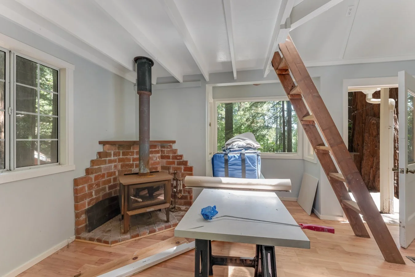 Room under renovation with a wood stove, large window showing trees outside, blue storage bin with laundry, and a homemade ladder leading to the ceiling.