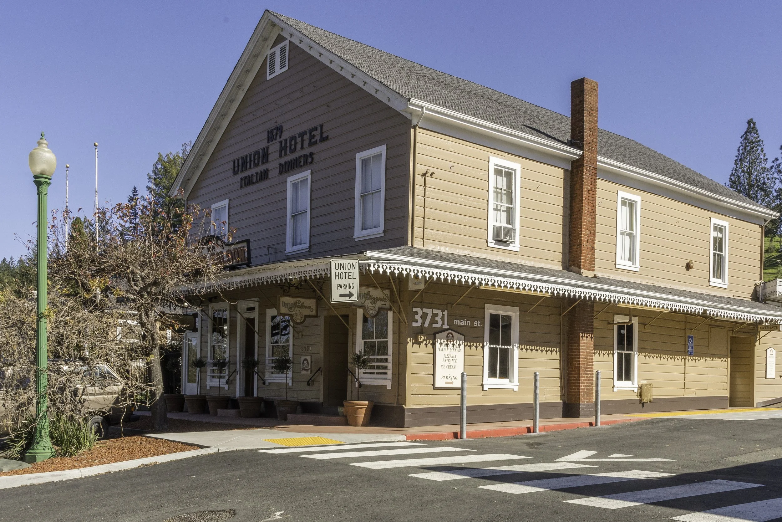 A two-story building with a sign reading 'Union Hotel' and 'Italian Dinners.' The building is painted a light peach color with white trim and has a red brick chimney. There is a parking sign and a crosswalk in front, with a green lamppost and leafles