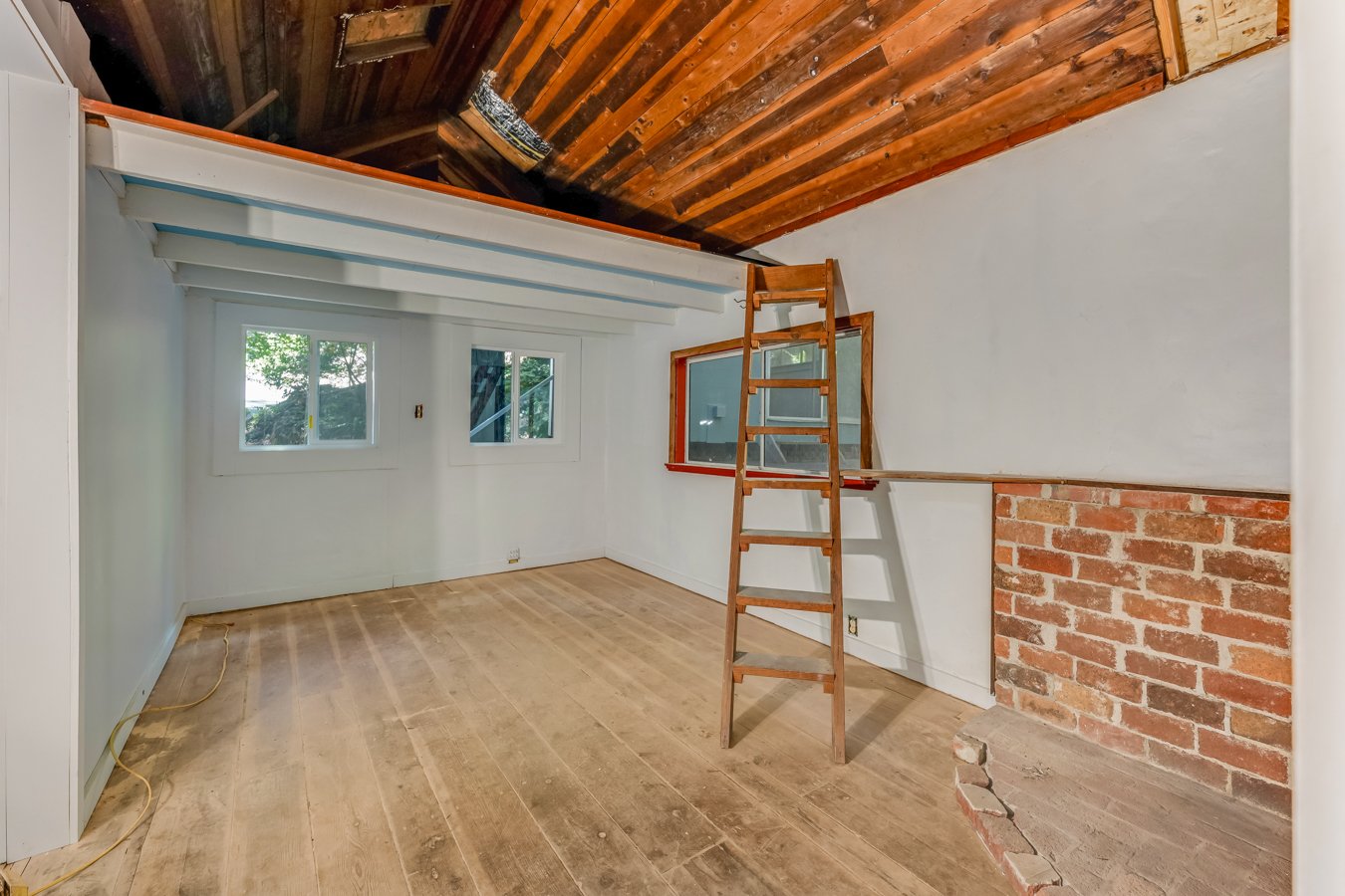 Empty room with hardwood floor, white walls, two windows, a brick feature wall, ceiling with exposed wood beams, and a ladder leading up to a loft area.