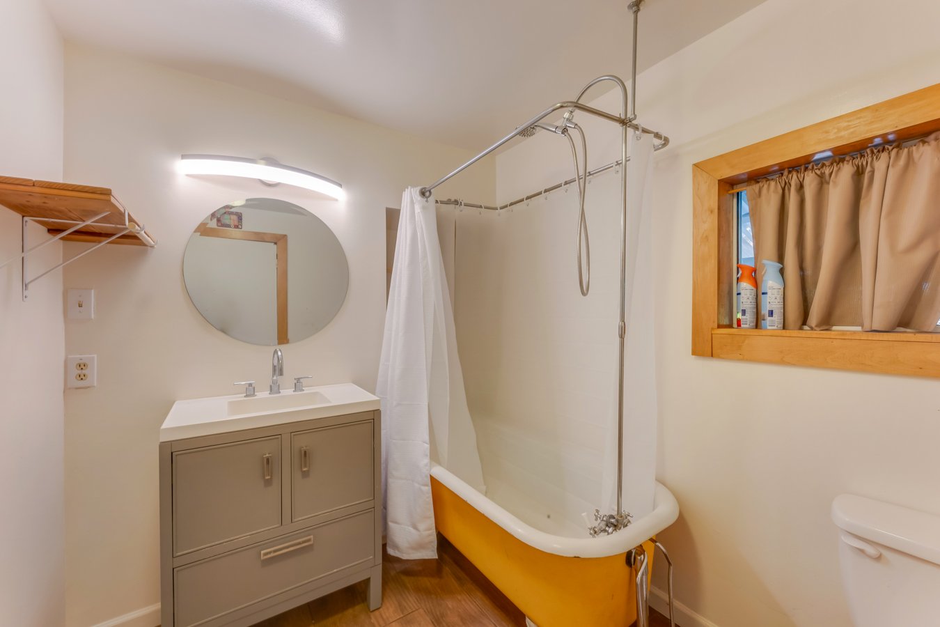 Small vintage bathroom with a yellow bathtub, a white curtain, a gray vanity with a sink, a round mirror, and a wood-framed window with beige curtains.