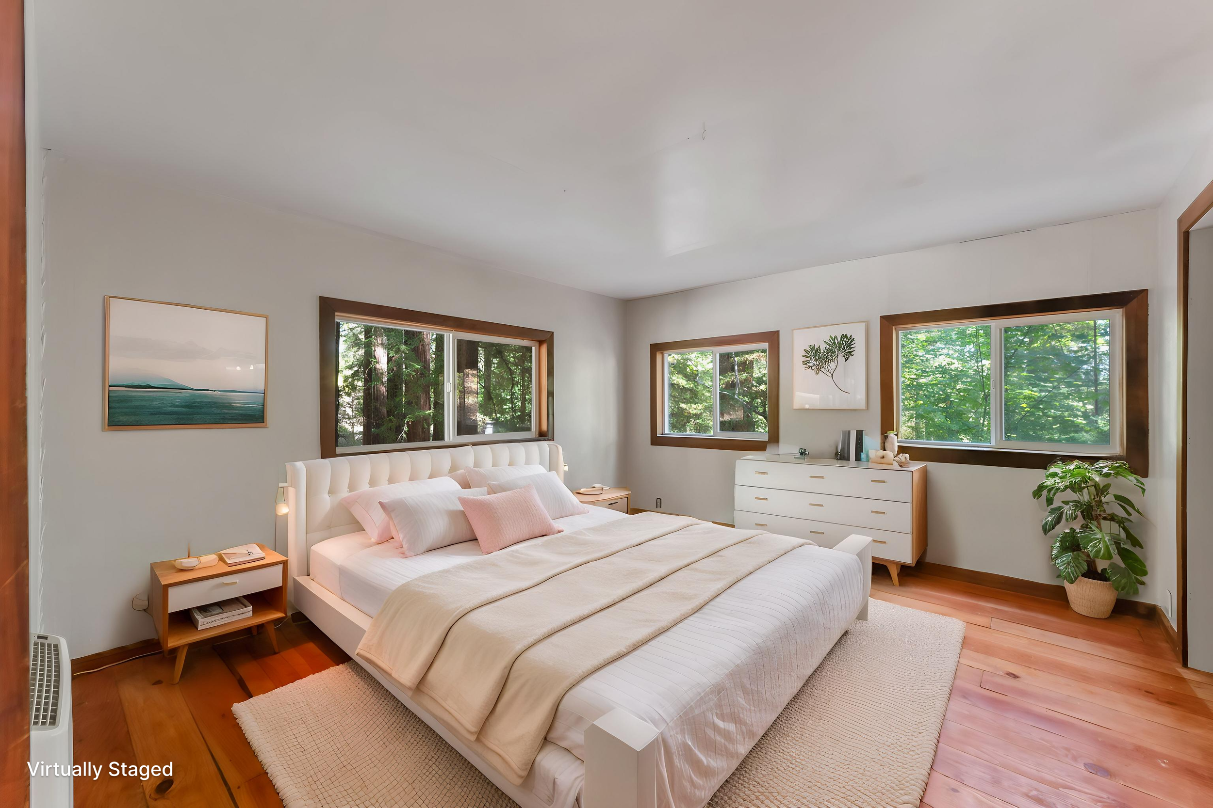 A cozy bedroom with a white bed featuring pink and beige pillows, wooden nightstands, and large windows showing green trees outside. Decorative art hangs on the light gray walls, and a large potted plant is in the corner. The room has warm wooden flo