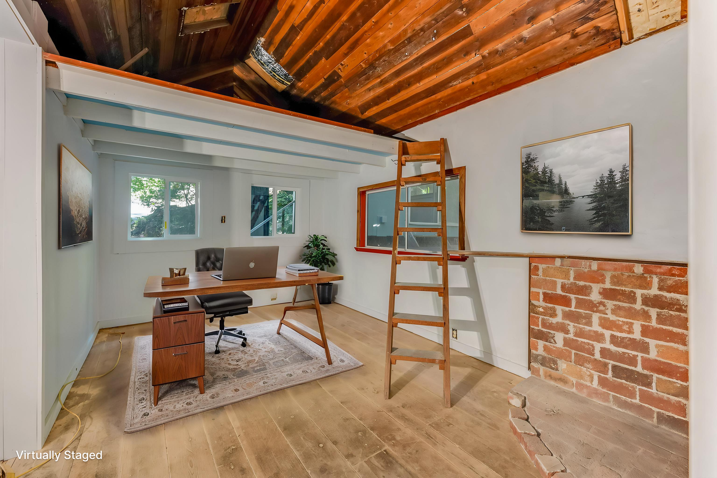 A home office with white walls, a wooden desk, a black office chair, two framed landscape photos, a potted plant, and a ceiling with wooden planks. A ladder leans against the wall, and part of a brick wall is visible on the right. Windows let in natu