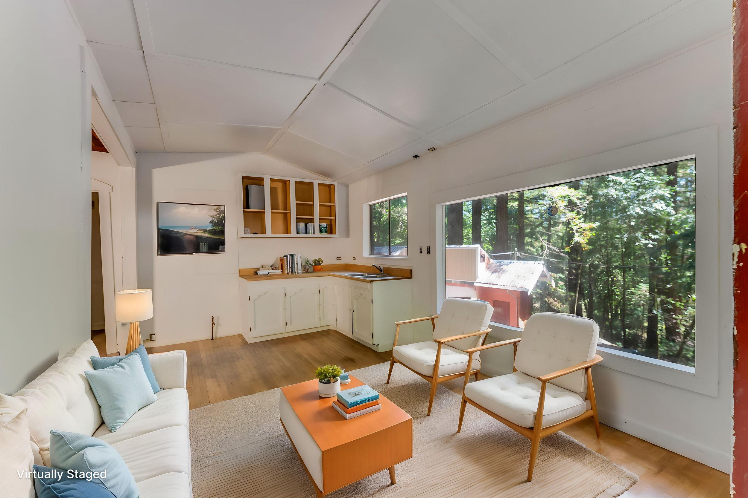 Living room with white couch and pillows, wooden chairs, coffee table with books, large windows showing a forest outside, and a kitchenette area with open shelves and a sink.