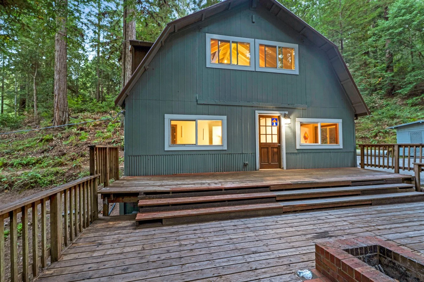 A two-story green wooden house with a wooden deck and stairs, surrounded by a forest of tall trees, with warm interior lights visible through the windows.