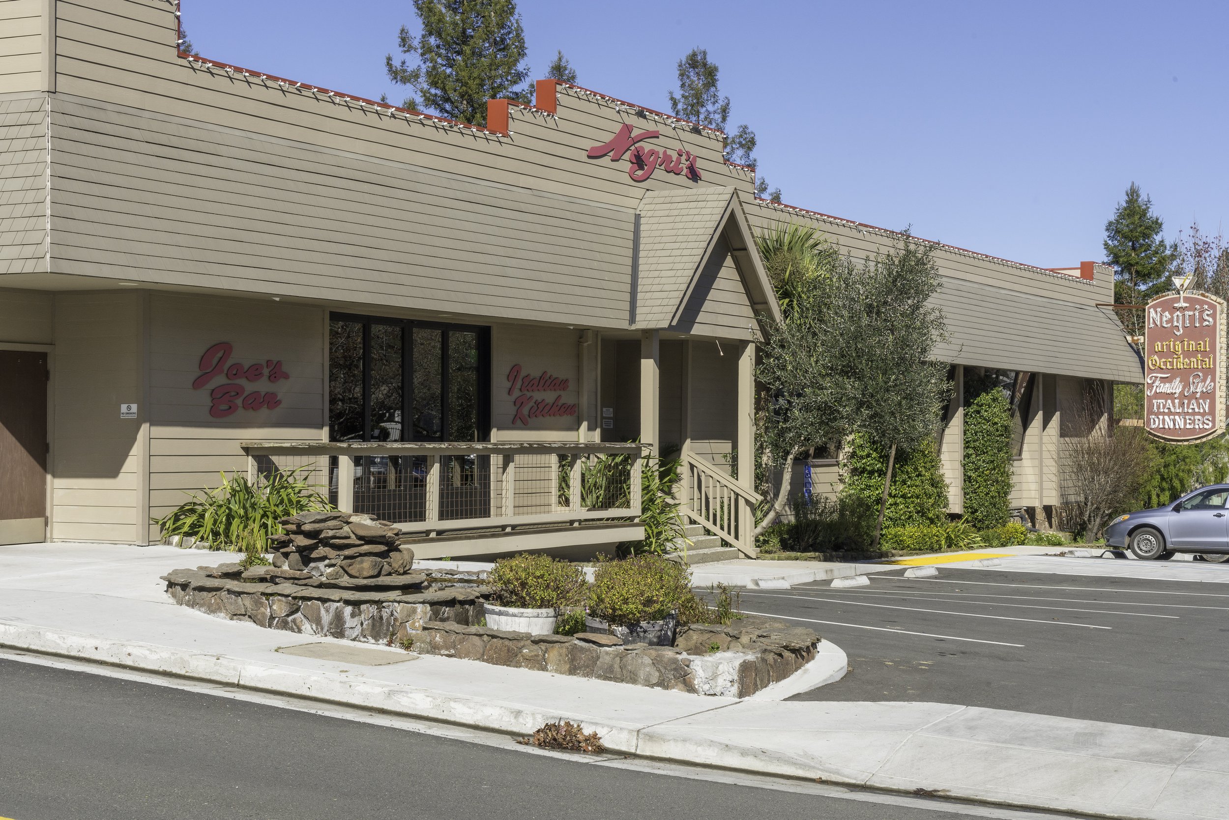 Exterior view of an Italian restaurant called Ngeri's, featuring a beige building with pink signage, a parking lot, and landscaped area with plants and trees.