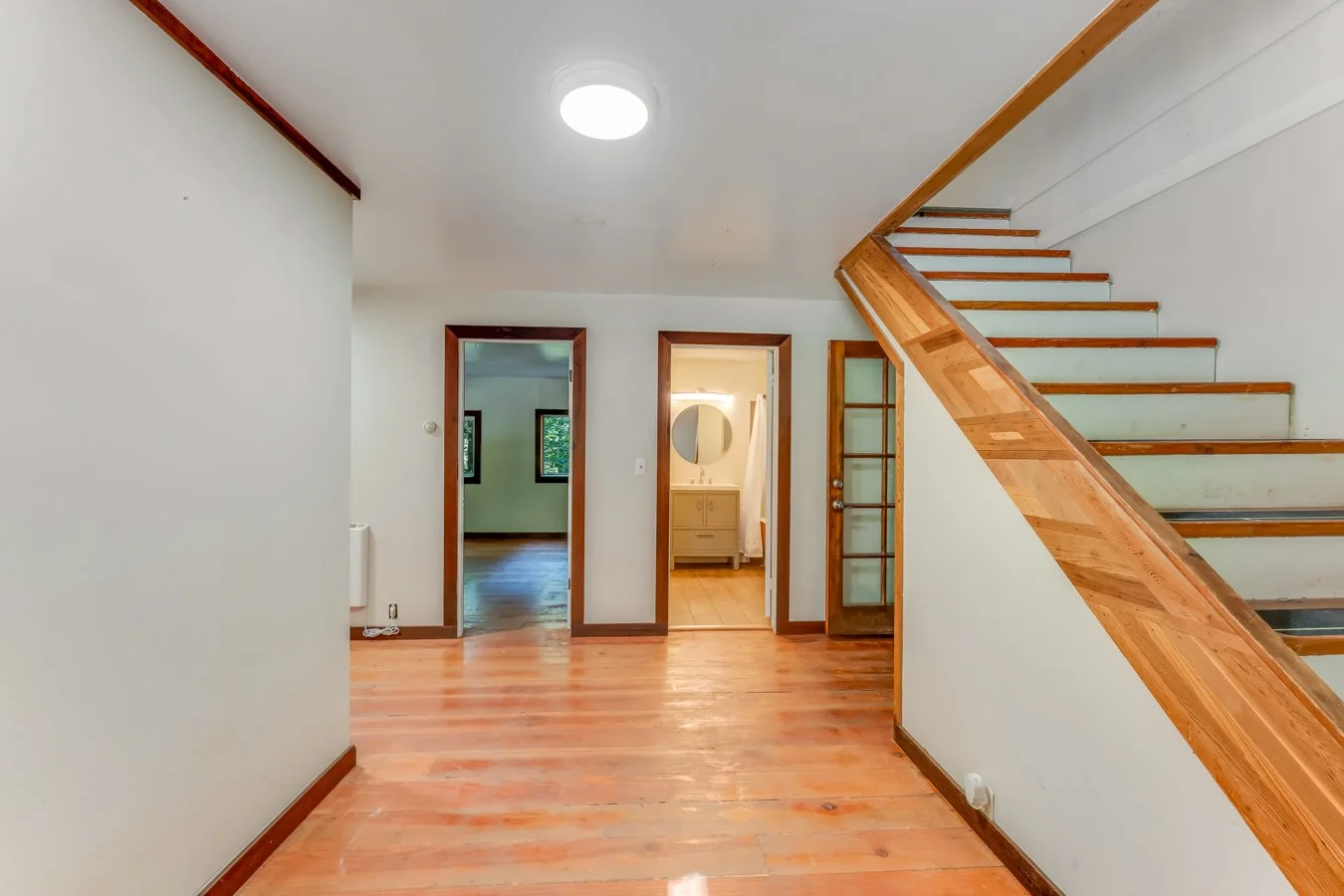 Interior view of a house showing a living area with hardwood floors, a staircase with wooden railing, three doorways leading to other rooms, and a ceiling light fixture.