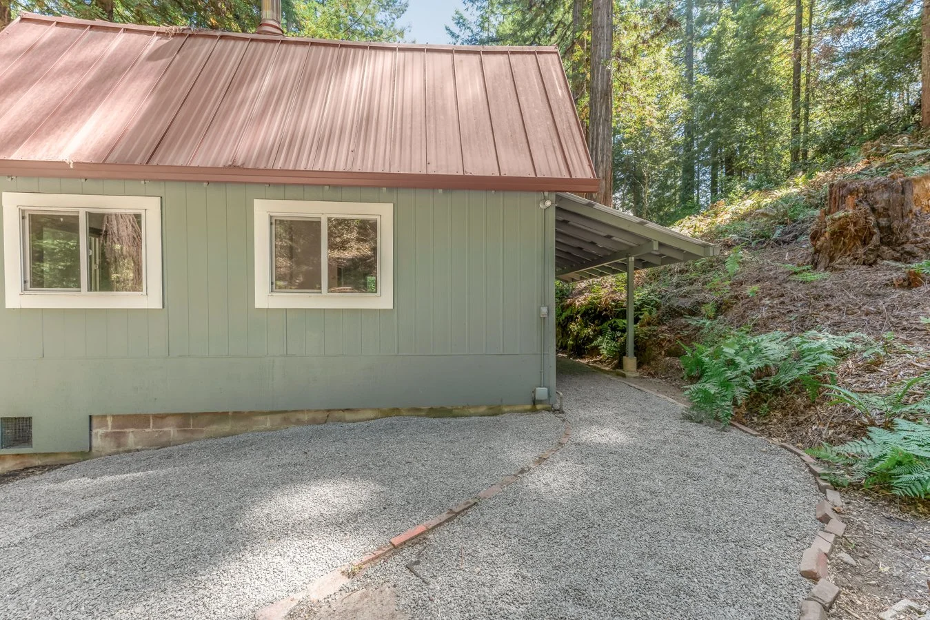 Exterior side view of a green house with a metal roof situated in a wooded area, with a gravel driveway and a small covered parking space supported by metal posts.