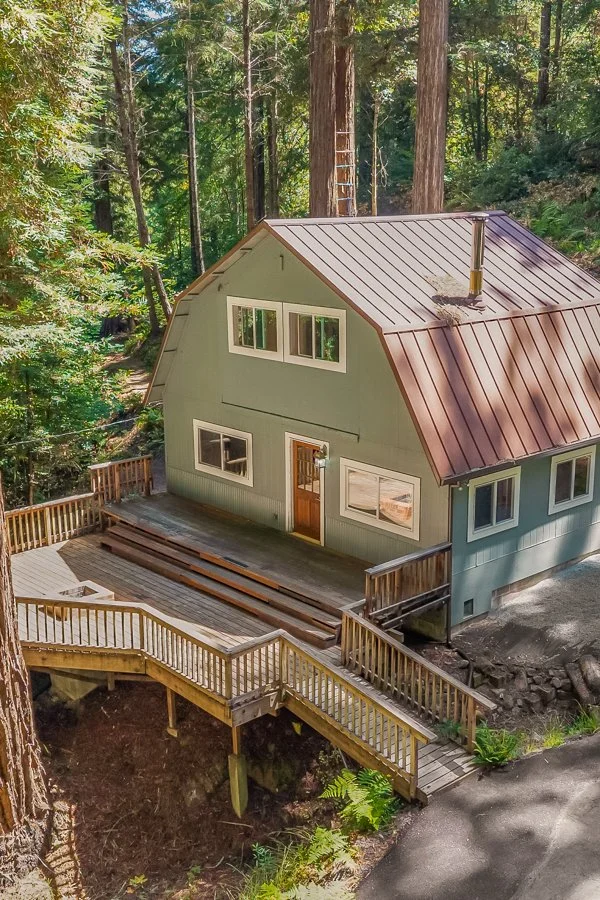 A renovated two-story house with a metal roof, surrounded by tall trees in a forested area. The house has green siding, multiple windows, and a front door with a small deck. There is a wooden staircase leading to the house and a large tree directly i