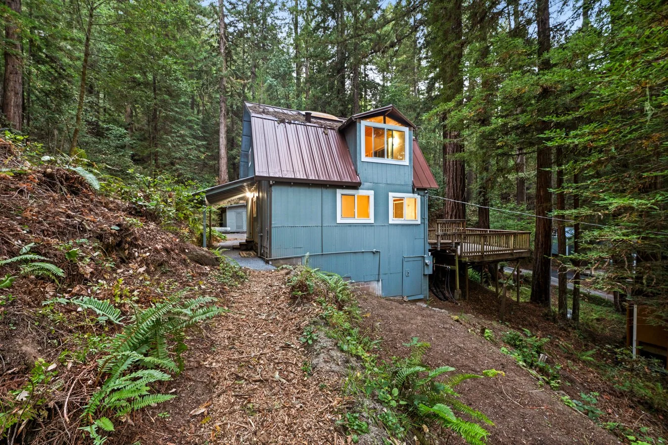 A blue house with a metal roof nestled among tall trees on a forested hillside, with a wooden deck and illuminated windows at dusk.