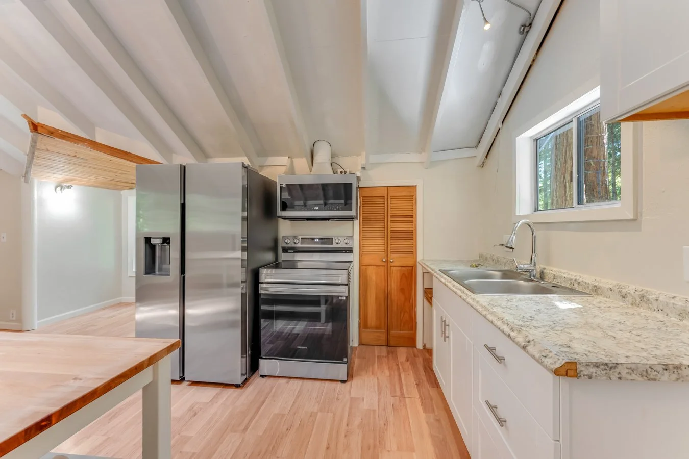 Kitchen with a granite countertop, white cabinets, a stainless steel double sink, a large window, a stainless steel refrigerator, a stove with microwave above, and wooden flooring.