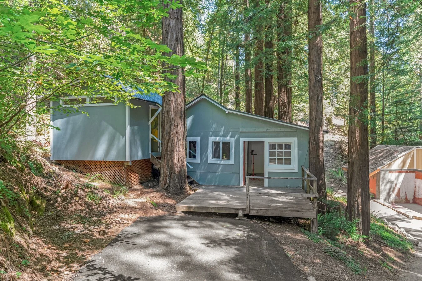 A house under construction in a forest surrounded by tall trees, with a wooden ramp leading to the entrance and a partially built foundation visible on the right side.