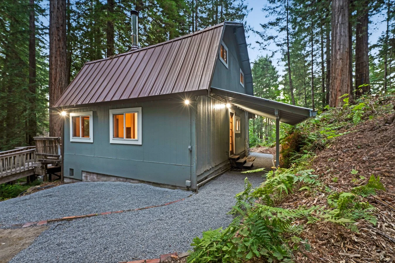 A cozy blue house with a metal roof in a forest, lit from inside, surrounded by tall trees and green foliage.