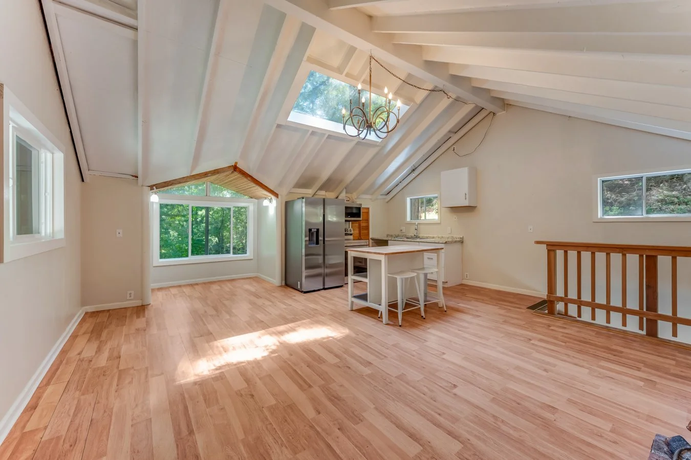 Open-concept living space with exposed vaulted ceiling, skylights, and hardwood flooring. Features a small kitchen area with a refrigerator, island, and white chairs, windows, and a chandelier.