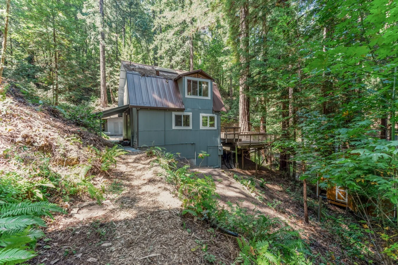 A small, two-story house with a metal roof is situated in a dense forest with tall trees and lush green foliage. The house has blue-gray siding, several windows, and a wooden balcony on the side. The ground around the house is sandy and slightly slop