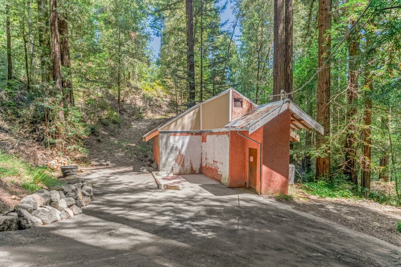 A small, unfinished building in a forest clearing, surrounded by tall trees, with a dirt driveway in front.