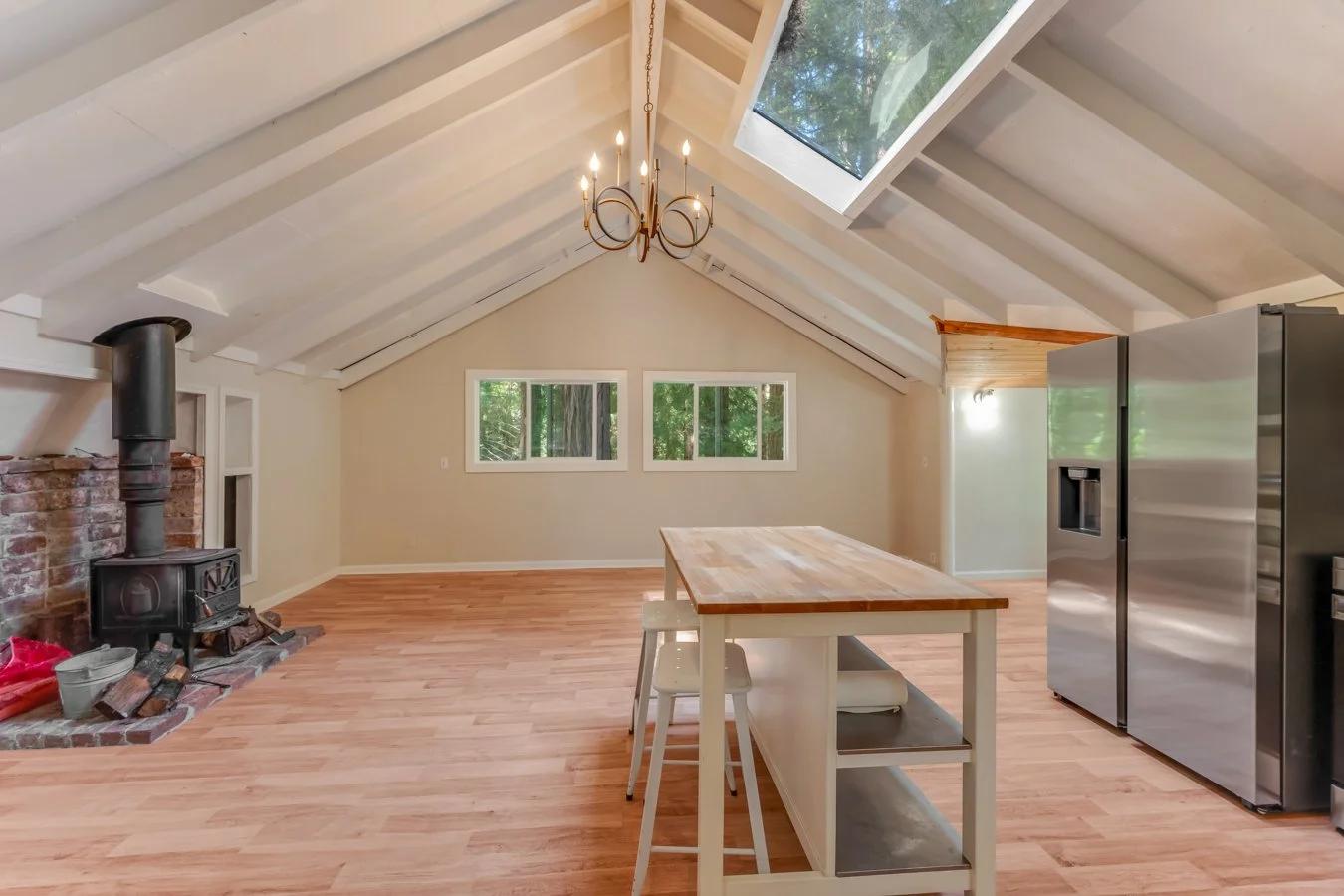 An open-concept kitchen or dining area with a vaulted ceiling, skylight, and wood flooring. Features include a black wood-burning stove near a brick hearth, a large stainless steel refrigerator, a wooden island with white base, and windows revealing 
