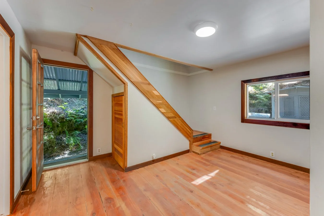 Empty room with wooden floors, large window, wooden door leading to outdoors, small wooden staircase, and sloped ceiling with wood accents.