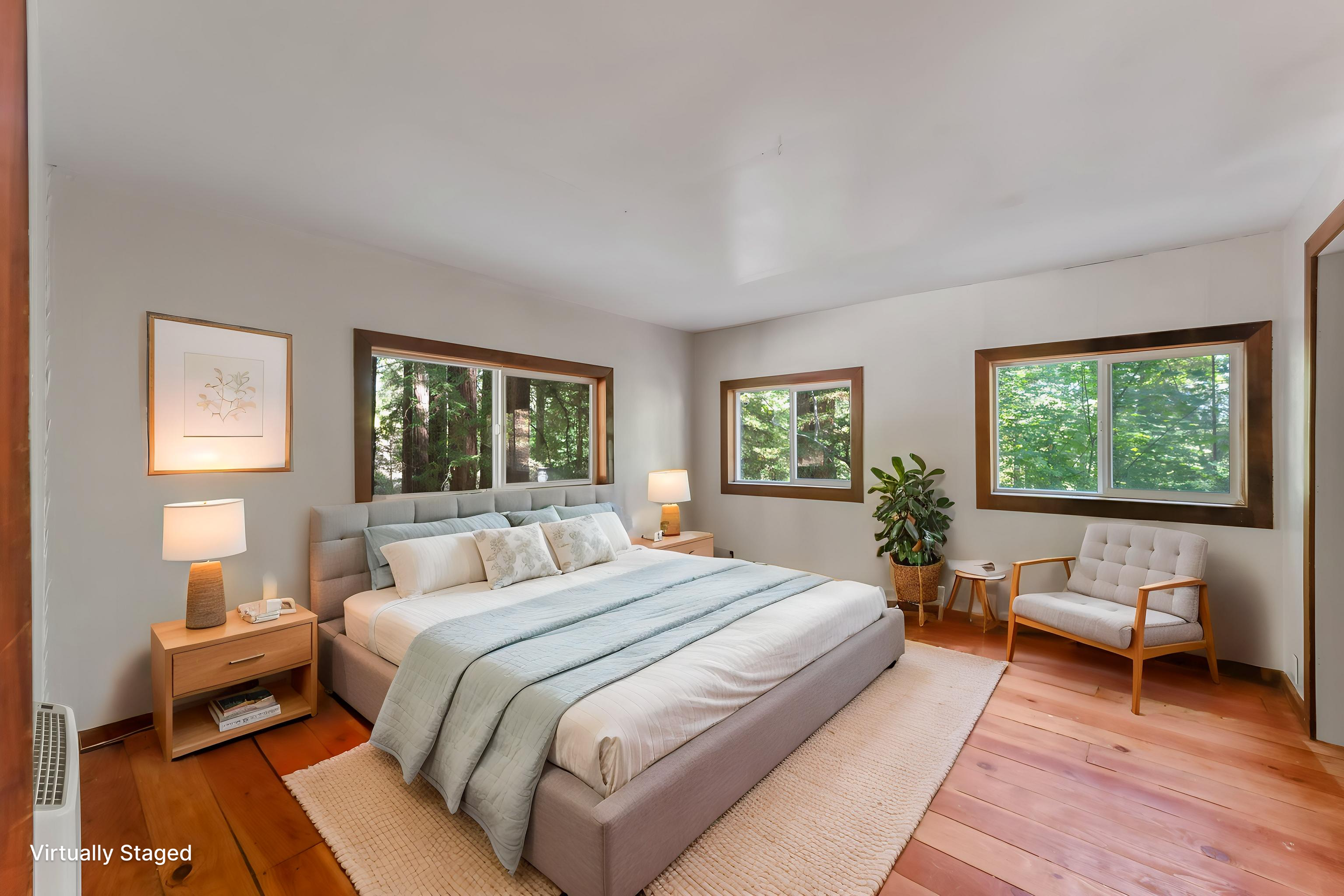 A bedroom with a large bed, nightstands with lamps, a framed wall art, and windows showing green trees outside.