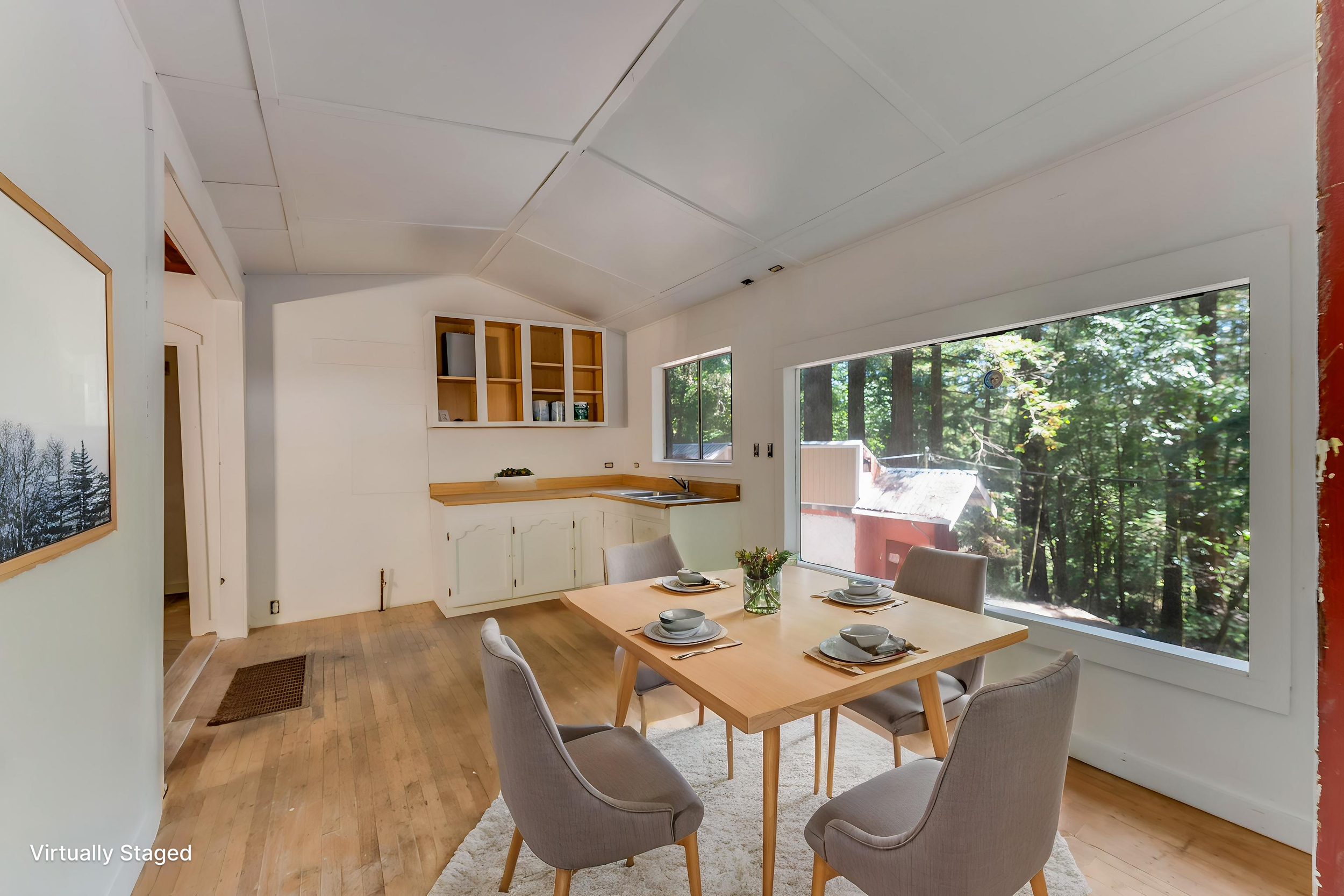 A dining area with a wooden table and six beige upholstered chairs, set with bowls, plates, and a glass vase with flowers, near large windows with a view of trees outside, in a bright room with white walls and ceiling, and wooden flooring.