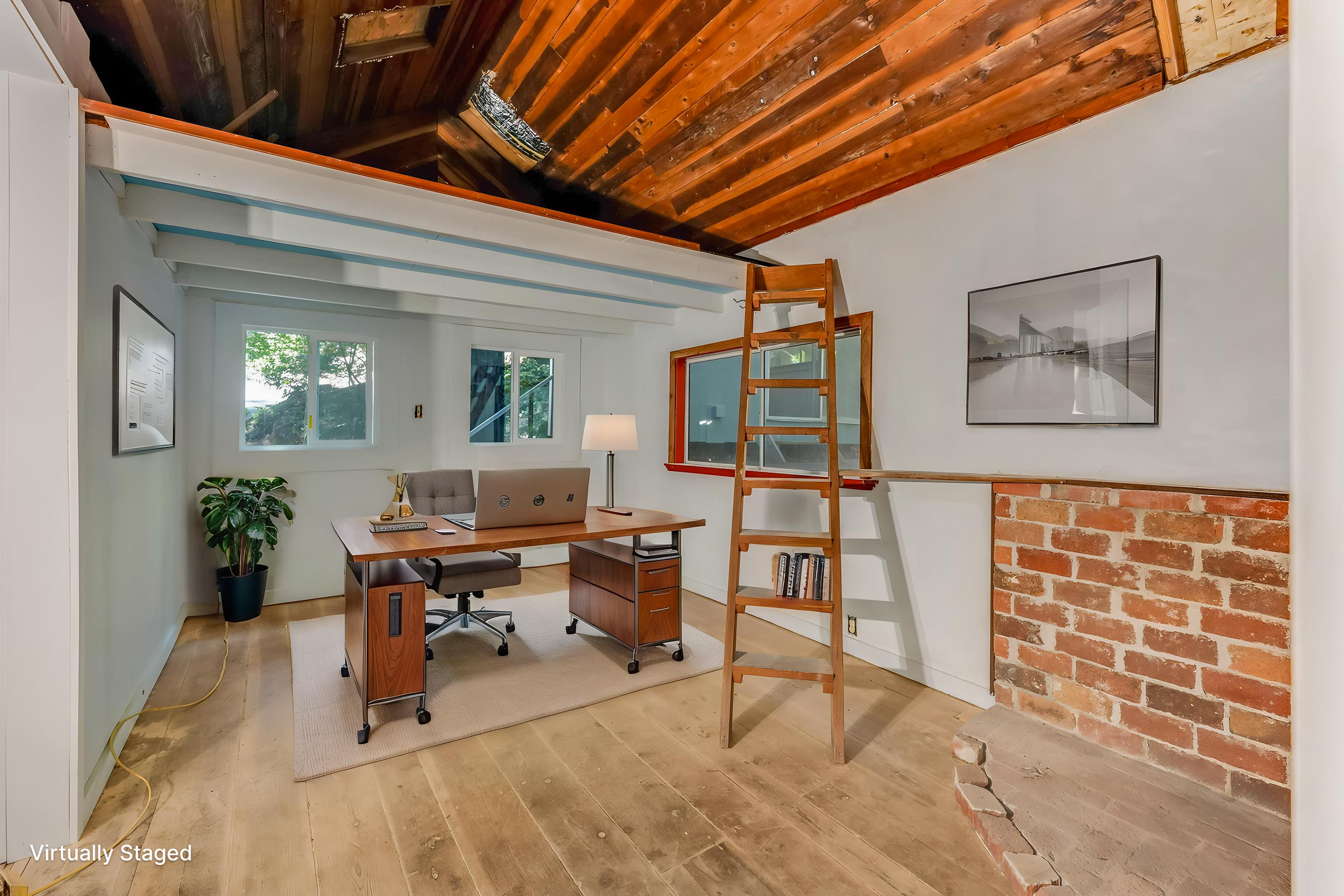 A home office with a wooden desk, gray office chair, potted plant, and desk lamp. Windows with trees outside, brick and white walls, with a ladder leading to a lofted space and a framed black-and-white photo on the wall.