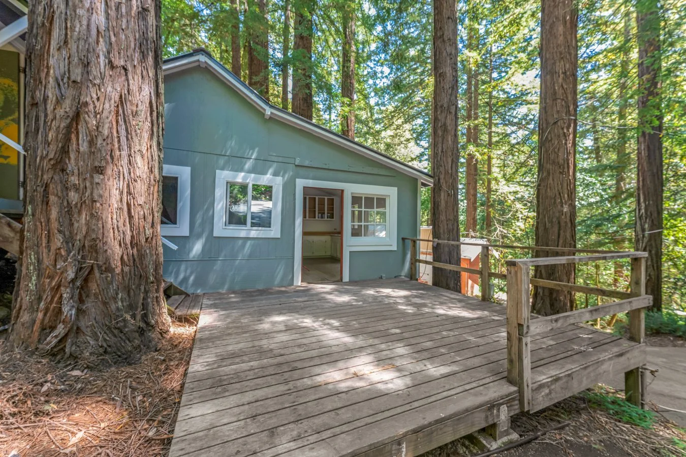 A small house or cabin with grayish-blue siding and white-framed windows, situated among tall trees in a forest. There is a wooden deck outside the house, partially shaded, with a railing around the edge.