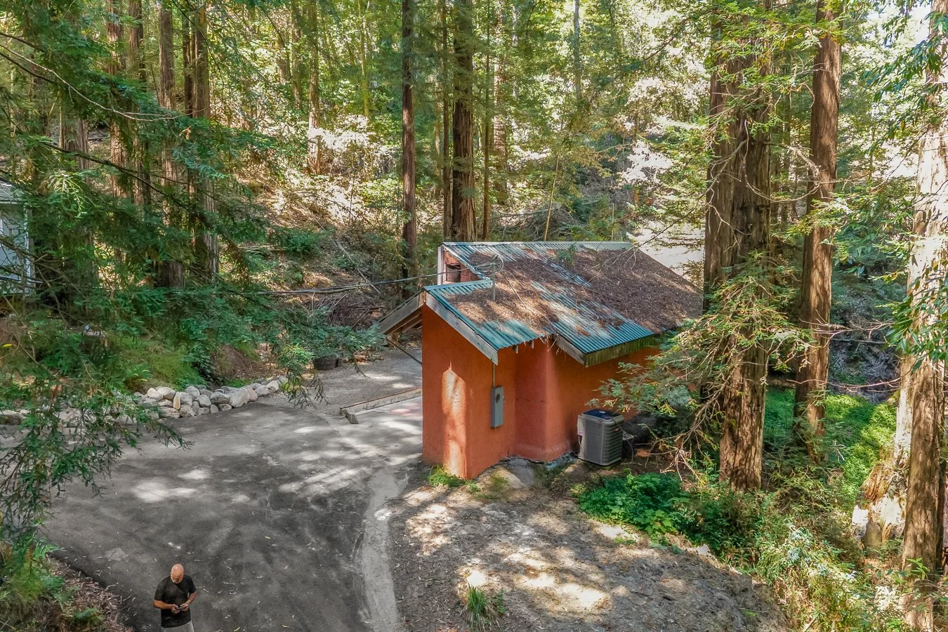 A small orange building with a rusted metal roof surrounded by tall trees in a forested area. A person is standing on the dirt ground, and an air conditioning unit is near the building.
