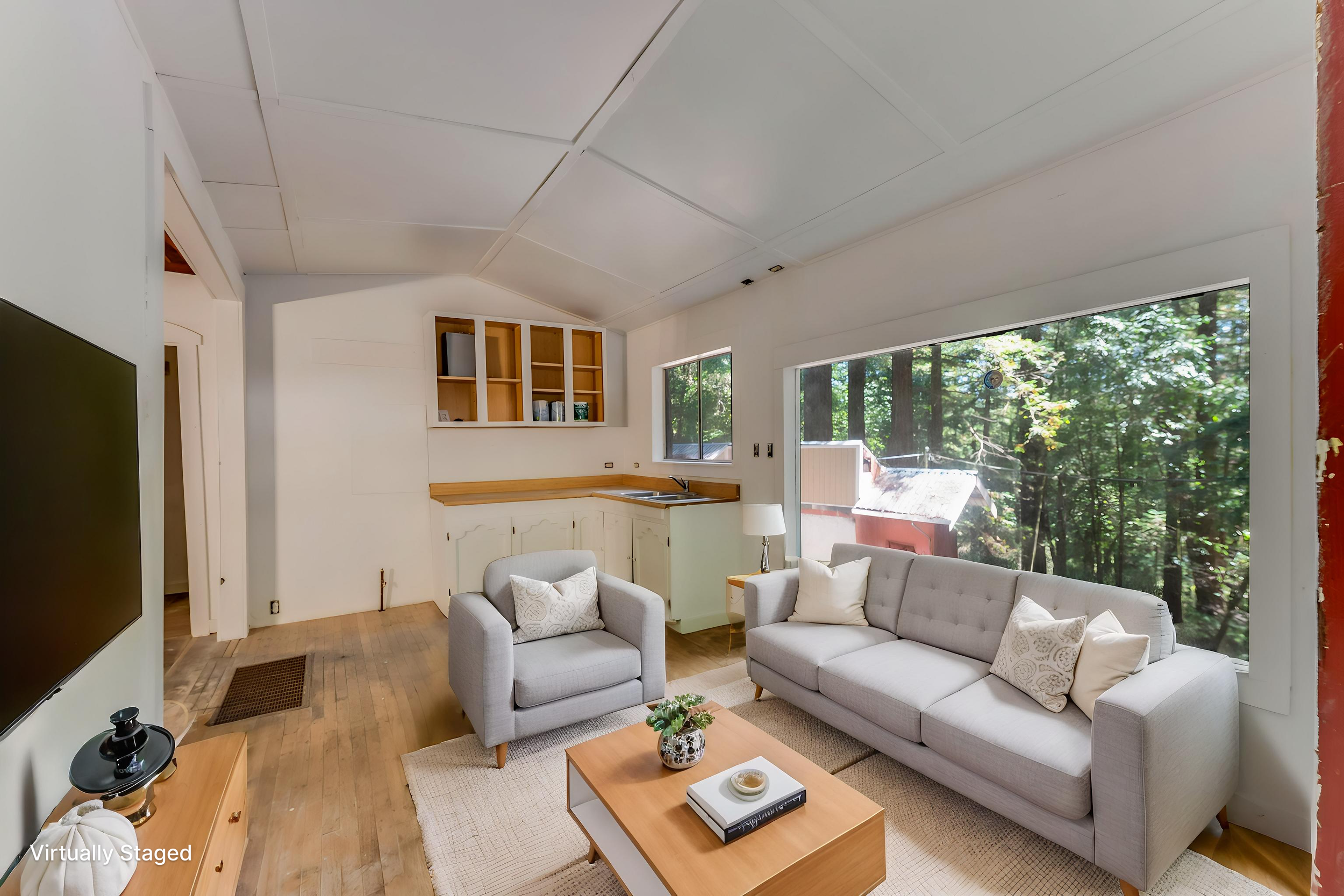 Living room with large windows showing a wooded backyard, light gray sofa with patterned pillows, matching armchair, wooden coffee table with book and plant, TV on wall, and unfinished kitchen cabinets