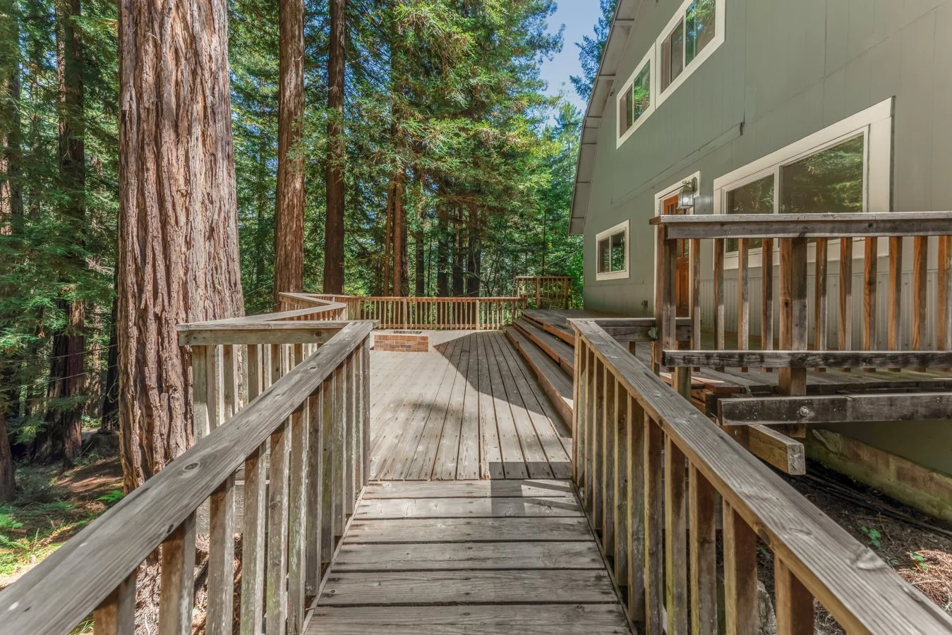 Wooden deck surrounding a house in a forested area with tall trees and green foliage.