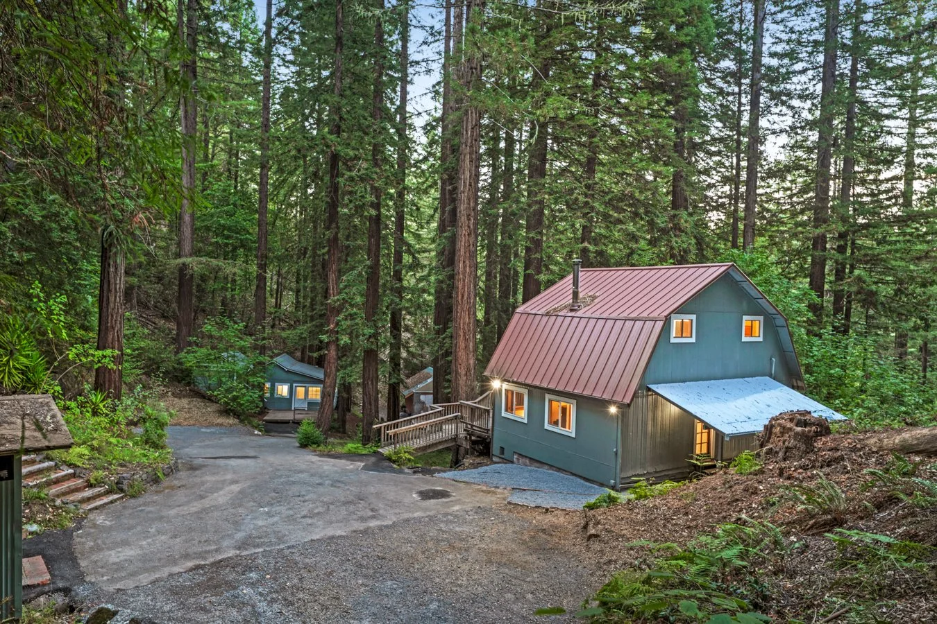 A cozy house with blue siding, situated in a wooded forest, with a gravel driveway and warm lights glowing from the windows.