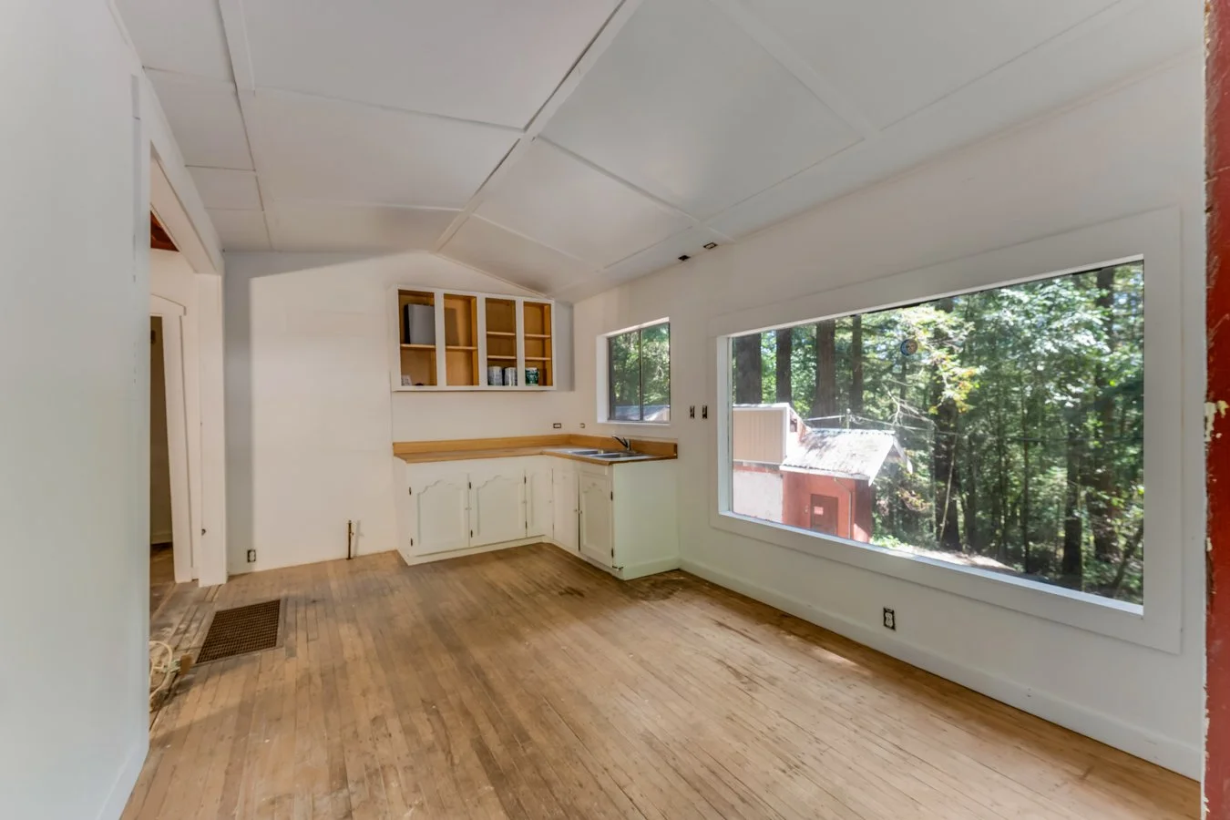 Empty kitchen area with large windows, white cabinets, wooden flooring, and open shelving, overlooking a wooded outdoor scene.