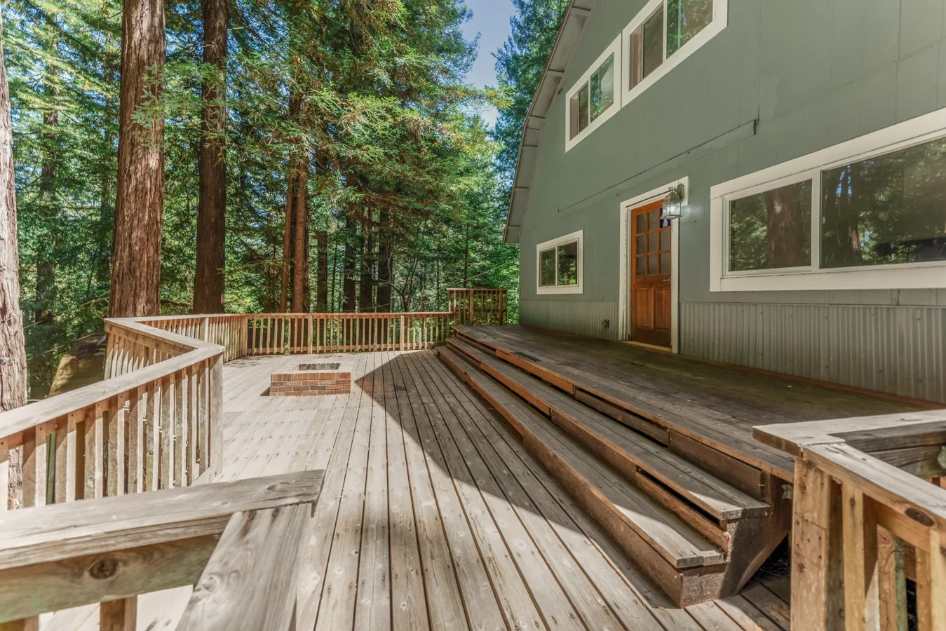 Wooden deck attached to a house in a forest, with a new railing and stairs, and tall trees surrounding the area.