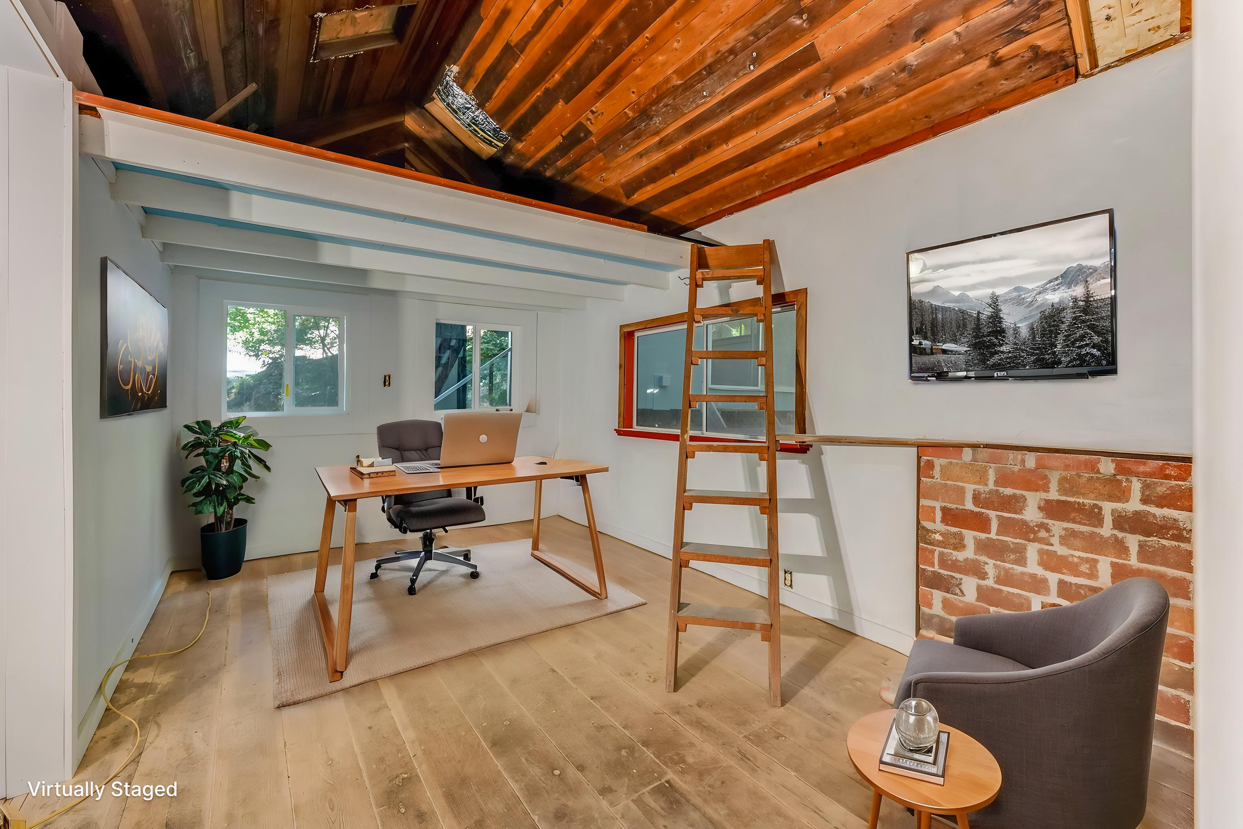Interior of a room with a wooden ceiling, white walls, and a brick accent wall. Furniture includes a wooden desk, office chair, armchair, and side table with a glass of water and a book. A ladder leans against the wall, and a large flat-screen TV is 