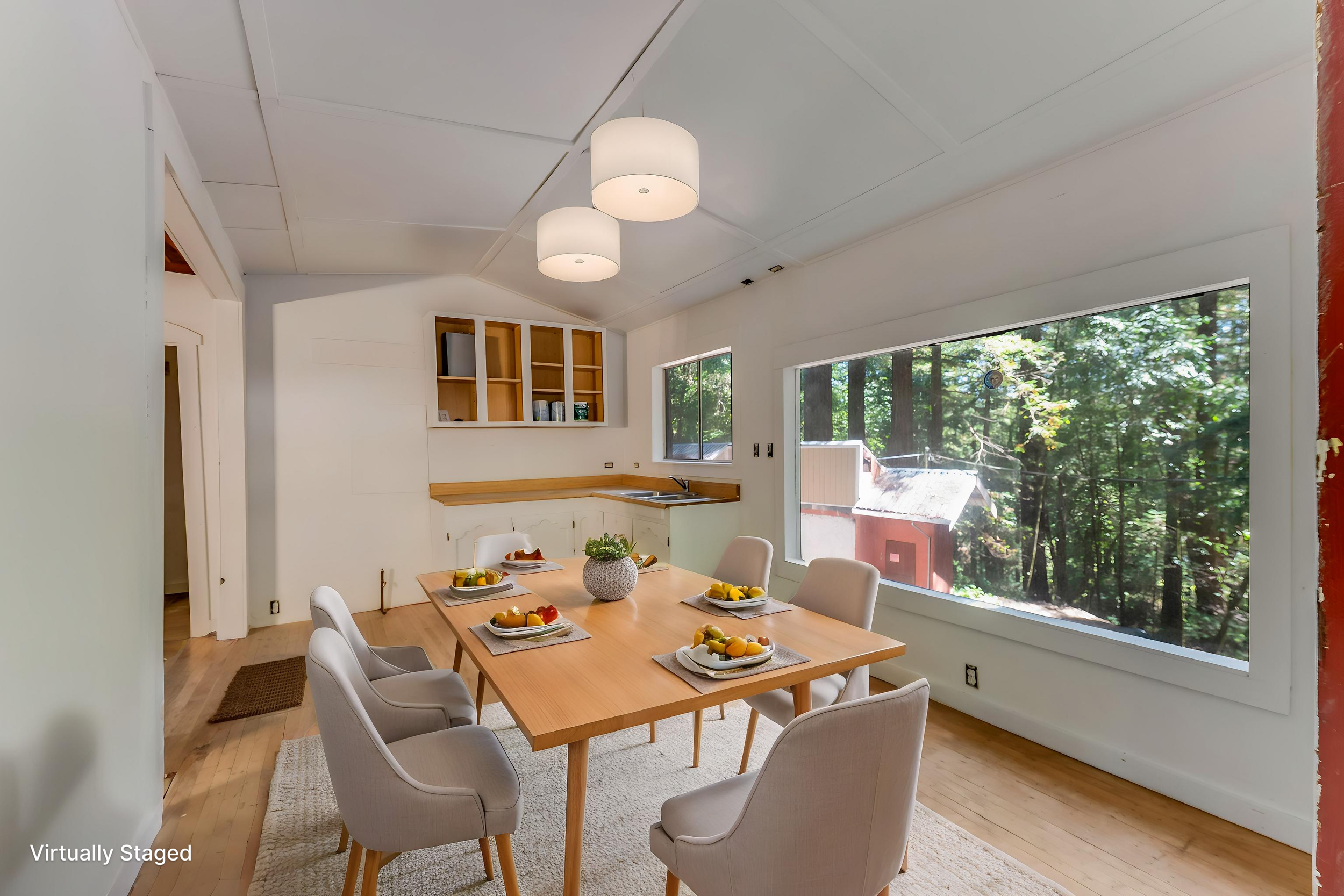 Dining room with a wooden table surrounded by white chairs, set with plates of fruit, in front of large windows showing trees outside. The ceiling has two modern white pendant lights, and there are built-in wooden shelves on the wall. The room is nat