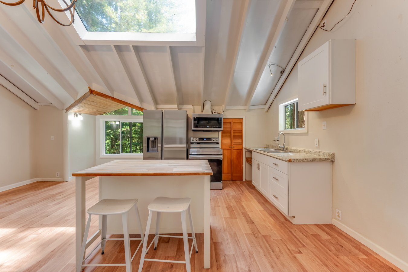 Interior of a kitchen with a sloped ceiling, light wood floors, white cabinets, granite countertops, stainless steel appliances including refrigerator and oven, small window above the sink, large window letting in natural light, and a white kitchen i
