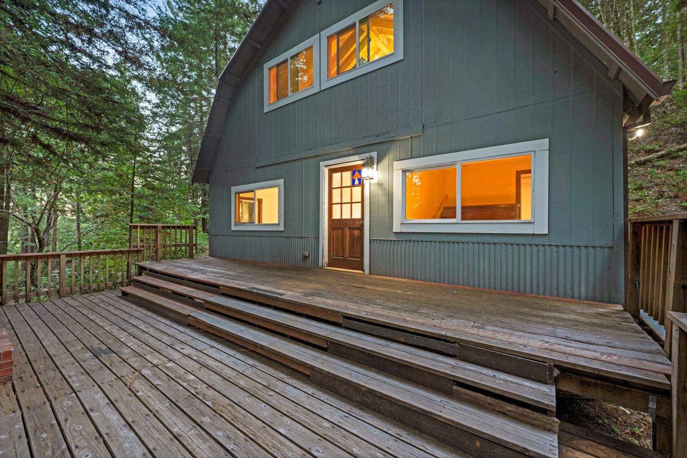 A two-story wooden house painted dark gray with white trim, surrounded by trees. It has a front porch with a wooden deck and steps, and lit warm interior windows visible through the glass.
